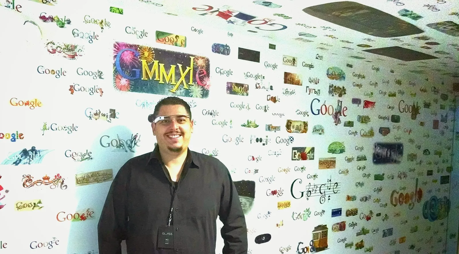 William wearing Google Glass in front of a wall of Google logos